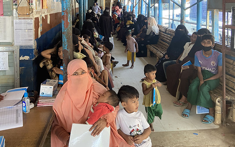 Salle d'attente d'une infrastructure de santé au Bangladesh. Au premier plan une mère voilée en orange portant dans ses bras son enfant, dans le fond de nombreuses mères et enfants