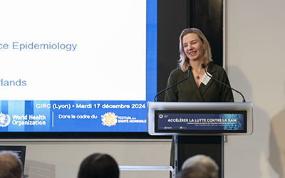 Carolien Ruesen stands at the lectern, smiling. In the foreground, the silhouettes of the audience.