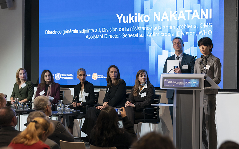 Five people are seated on a stage, a man and a woman stand in front of a desk, behind them a large blue screen.