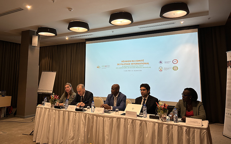 In a meeting room, five people are seated in a line on a table in front of a screen projecting the name of the International Steering Committee.